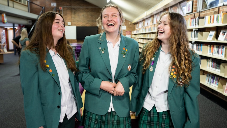 Bryndice, Harriet and Abi wearing green formal school jackets adorned with badges, standing together and laughing in a library.