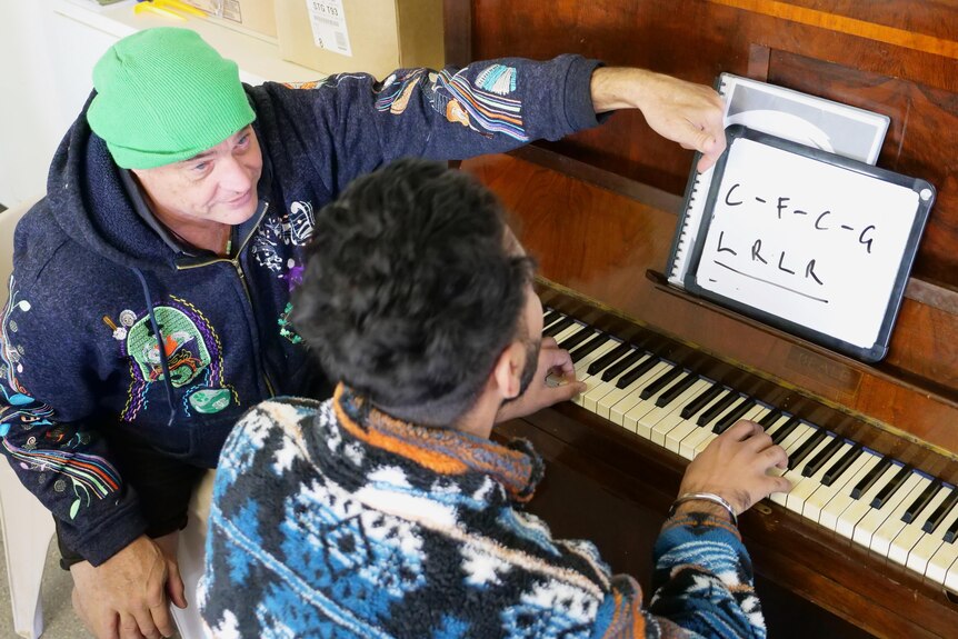 Mark McGurgan pointing to a whiteboard sitting on a piano that Mani Suhi is sitting at. 