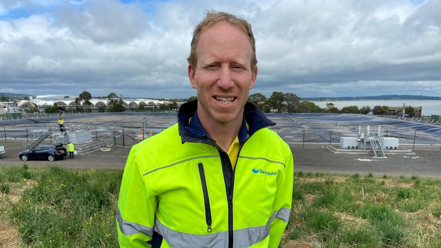 A man with short blond hair, wearing a fluorescent yellow jacket, stands outside in a field.