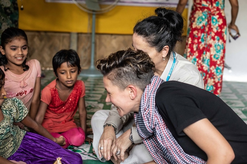 A woman sits on the ground laughing with three young girls and another woman.