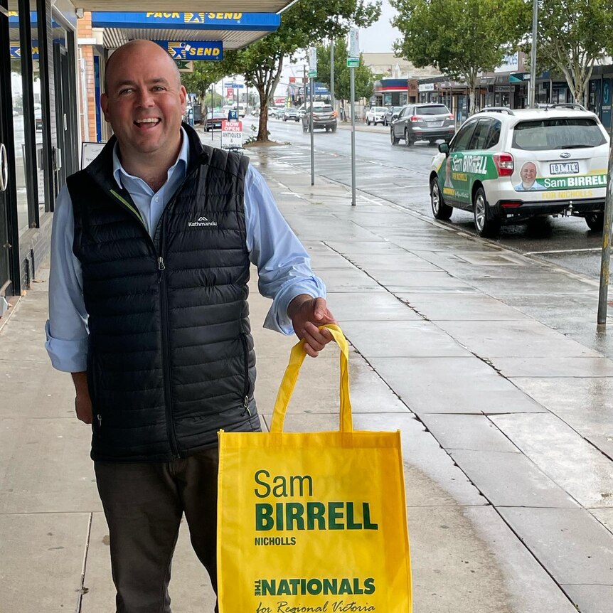 Sam Birrell smiles for a photo on a street while holding a yellow shopping bag branded with his campaign.