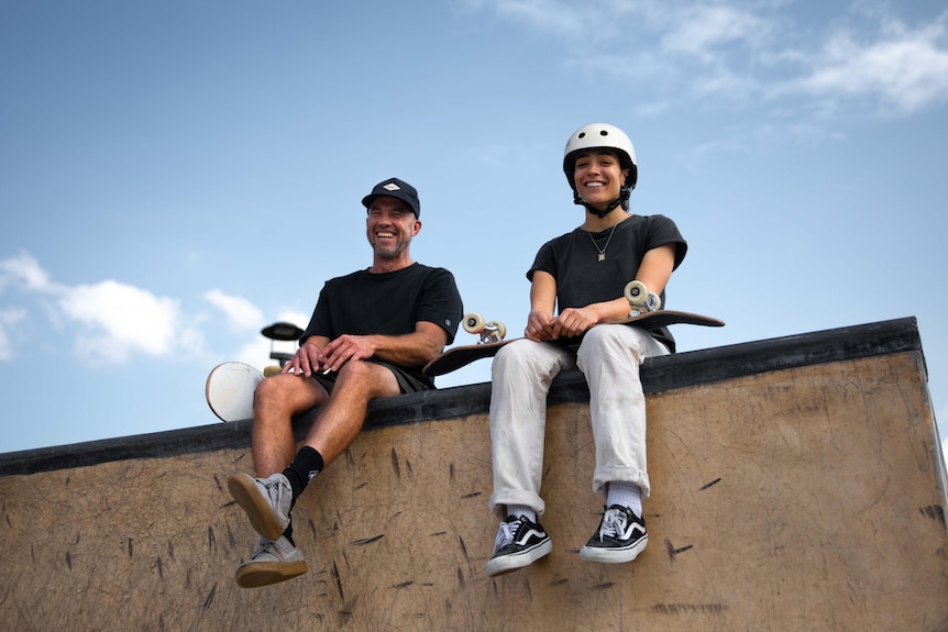 A man and a young woman sit laughing atop a skate park bowl.