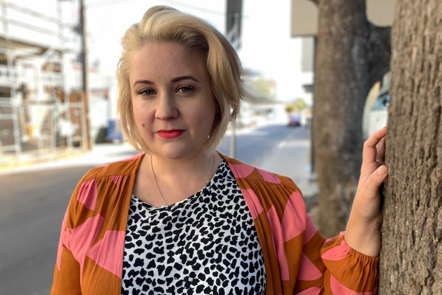 Chrissie Maus stands in a street leaning against a tree.