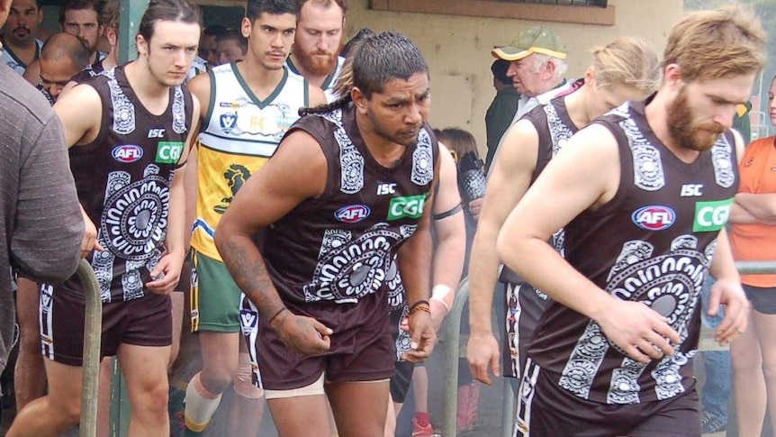 A heavyset country AFL player runs out of the shed for a match. He is wearing an ankle bracelet.