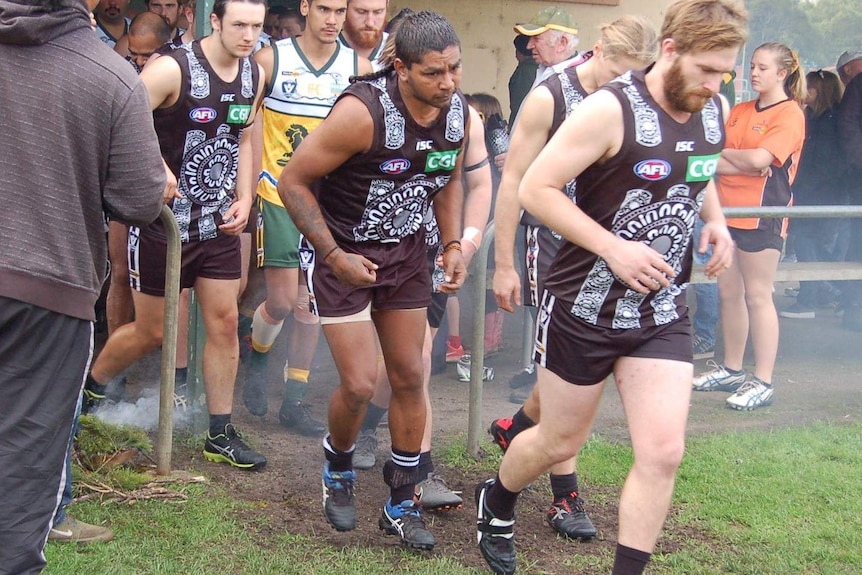 A heavyset country AFL player runs out of the shed for a match. He is wearing an ankle bracelet.