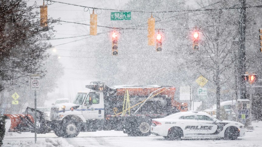 A snow plough and a Carrboro police vehicle pass each other in snowy conditions at a set of traffic lights.