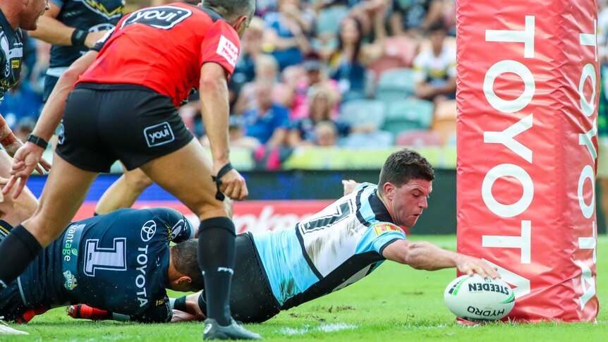 An NRL player avoids a tackle to reach out to plant the ball next to the posts for a try.