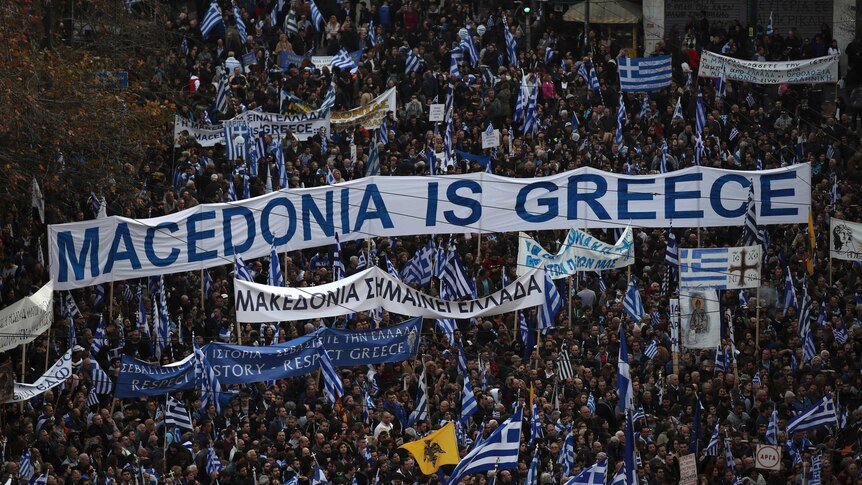 Thousands of protestors gather during a rally, with many carrying banners. One banner says 'Macedonia is Greece'.