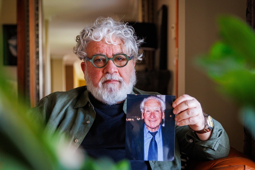 A man, sitting inside a house, holds a photo of his elderly father.