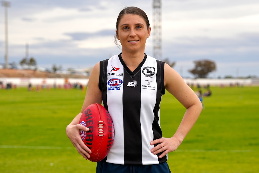 A white womanwith brown hair wearing a black and white football guernsey and holding a red footy