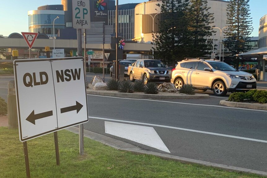 A line of cars are stopped on the street next to a sign that says "QLD/NSW".