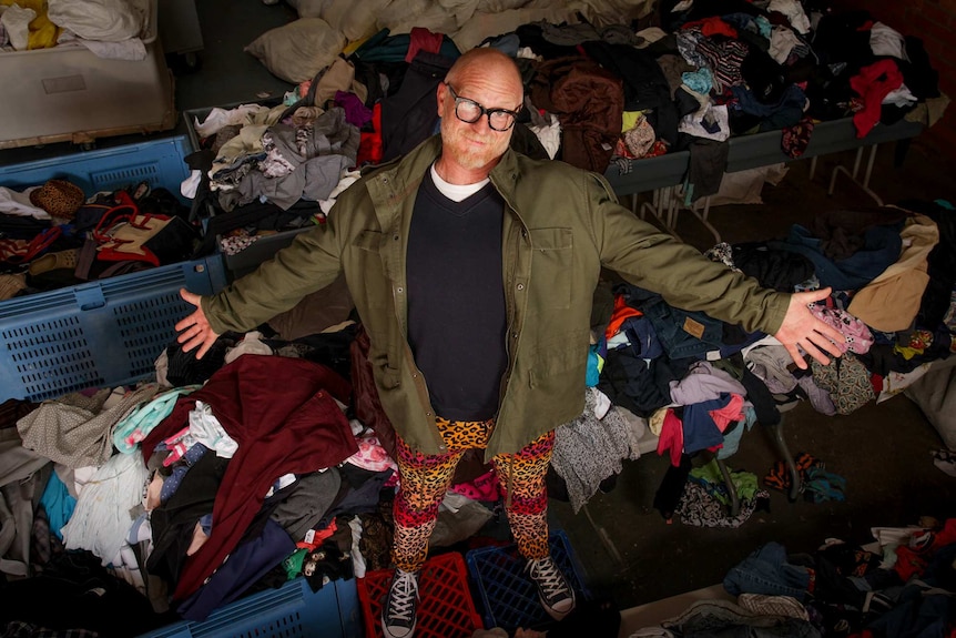 Man standing in the middle of piles of second hand clothing with his arms outstretched