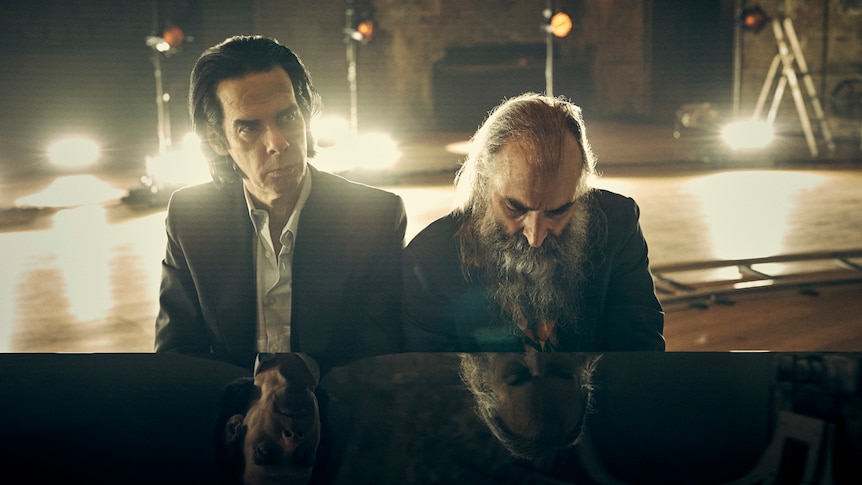 Nick Cave and Warren Ellis, two middle-aged men, sit at a piano together, Cave looking into the distance, Ellis at the keys