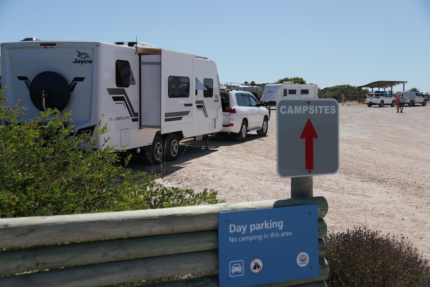 A sign that reads "campsites" in front of a gravel road with some caravans on it.