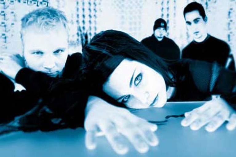 A band pose for a publicity photo, with the female lead singer placing her face on a table and reaching forward.