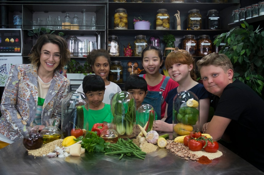 Alice stands at a kitchen bench with six children and lots of fresh food.