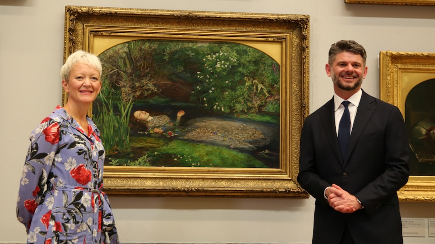 Maria Balshaw (left) in floral dress and trainers and Nick Mitzevich (right) in black suit, on either side of painting.