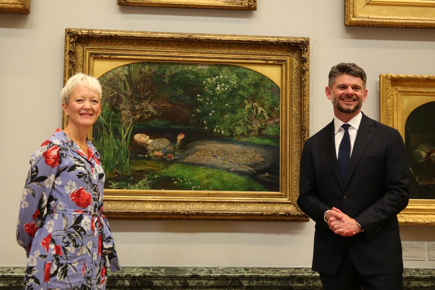 Maria Balshaw (left) in floral dress and trainers and Nick Mitzevich (right) in black suit, on either side of painting.