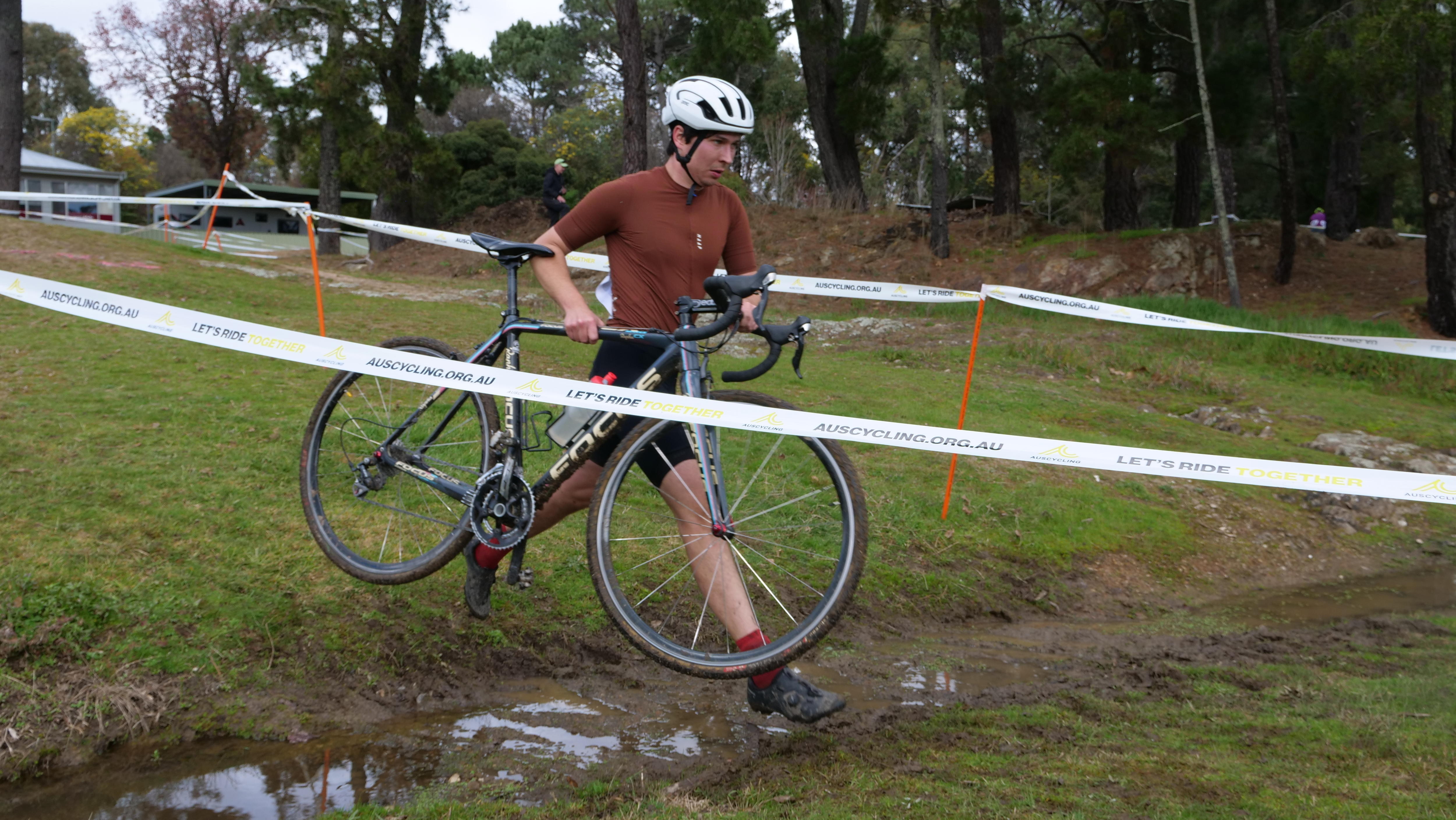 A male cyclist carries his bike across a watery and muddy ditch in the middle of the race course.