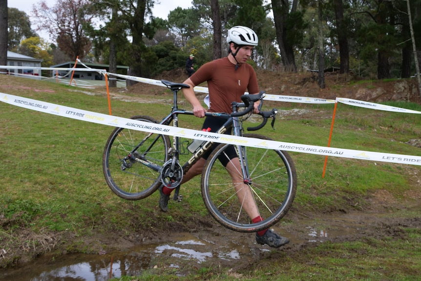 A male cyclist carries his bike across a watery and muddy ditch in the middle of the race course.
