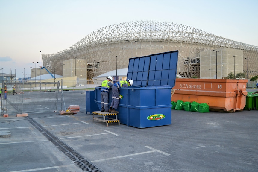 A man leans over a large blue, metal bin in front of a soccer stadium