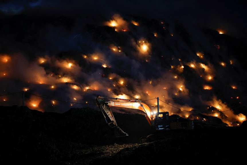 An excavator tries to contain a fire as smoke rises from burning garbage.
