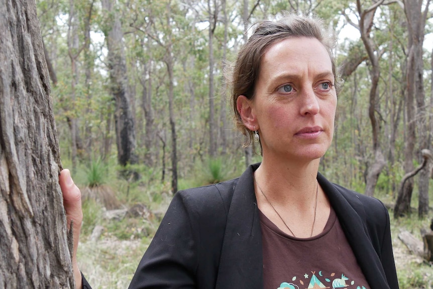 A woman with a t-shirt reading "forests for life" stands next to a tree in a bushland setting.