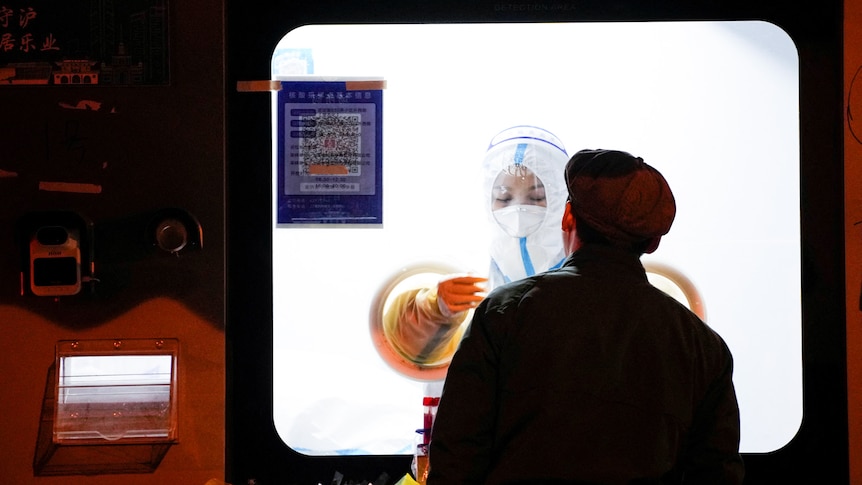 A medical worker in a protective suit wears gloves as they hold a swab up to a man wearing a coat and hat.