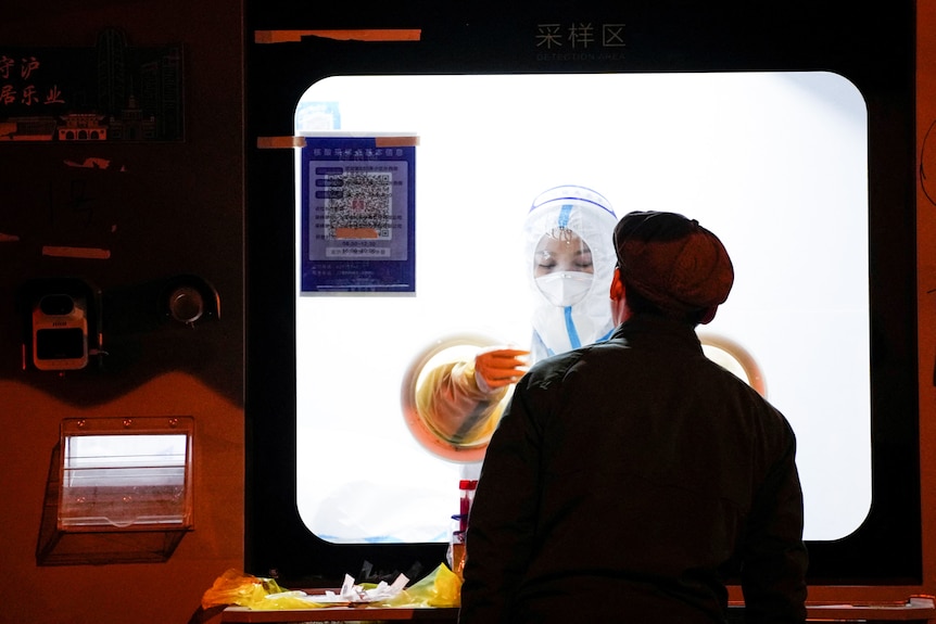 A medical worker in a protective suit wears gloves as they hold a swab up to a man wearing a coat and hat.