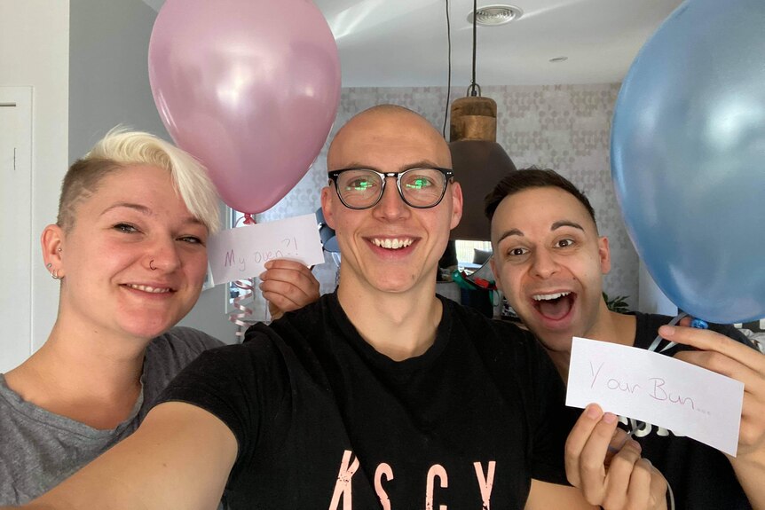 Woman and two men take a selfie during surrogacy proposal, they hold a pink and blue balloon and notes saying: Your bun, my oven