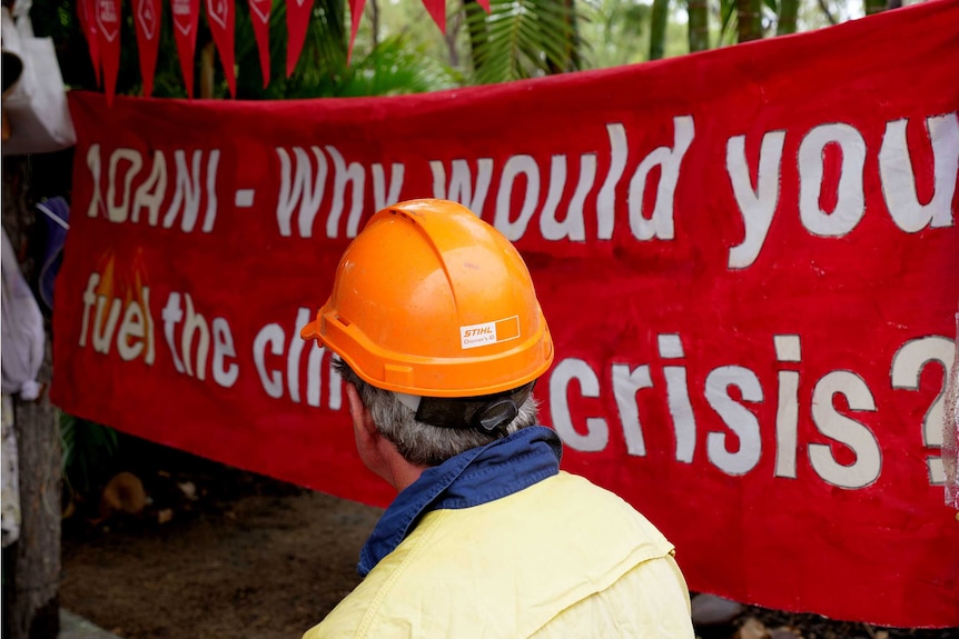 A man wearing an orange hard hat is looking at a red painted sign about Adani.
