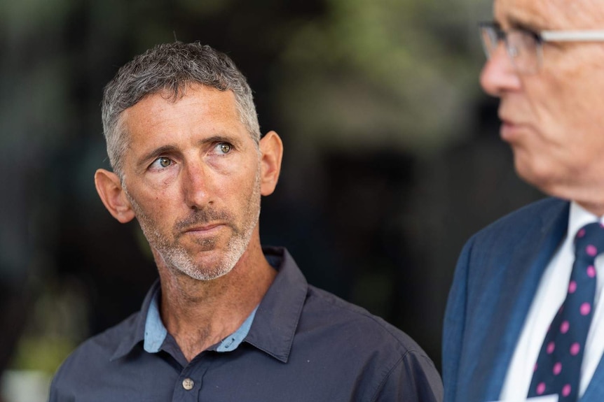 Aaron Cockman wearing a dark blue shirt in focus with John Quigley out of focus on the side.
