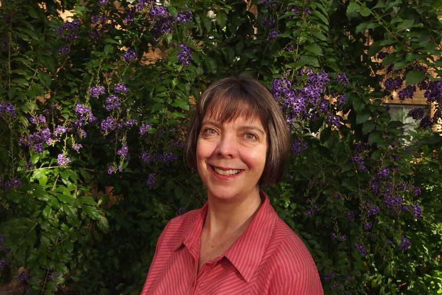 portrait of Caucasian woman with short brown bob wearing pink blouse smiling in front of purple leafy flowers