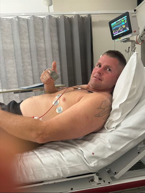 A man lies in hospital with wires attached to his chest. He gives a thumbs up to the camera