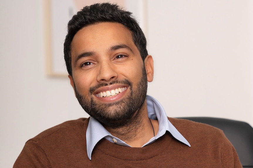 A bearded man wearing a brown jumper, smiling.