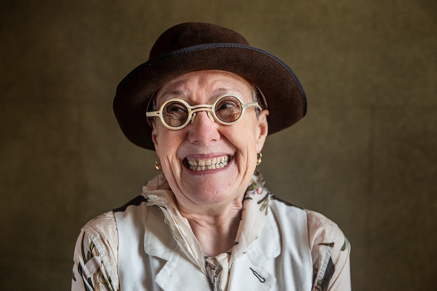 58-year-old woman with short hair, round-framed white glasses and dark  brown felt hat, smiling and looking away from camera.