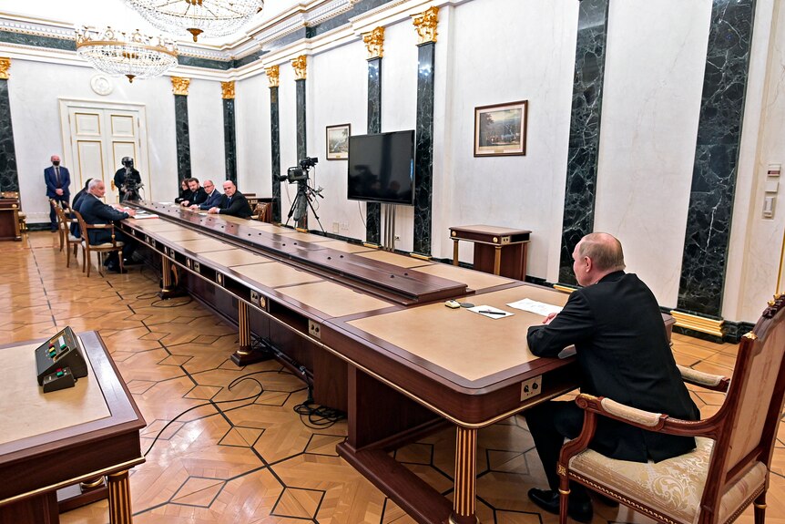 Putin, a man in a dark suit, sits at the head of a long table at which several people are sitting at the opposite end