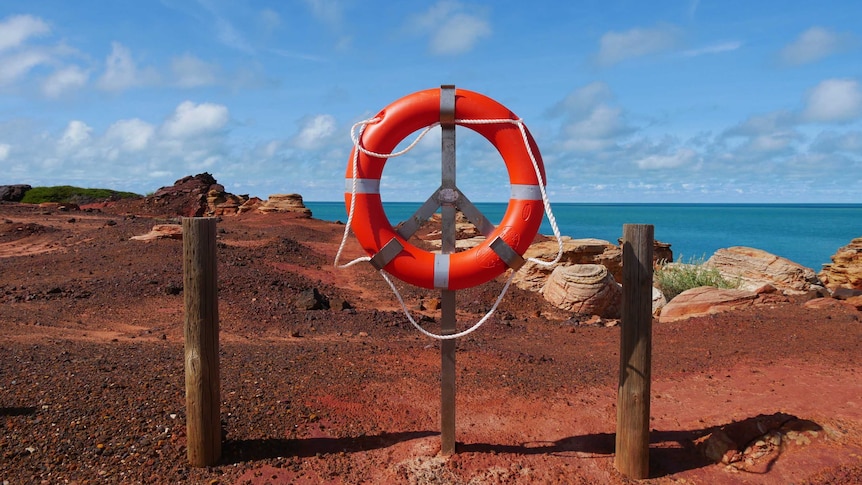 A life ring mounted on a steel pole in red dirt atop a cliff with the blue ocean behind it and clouds in the sky