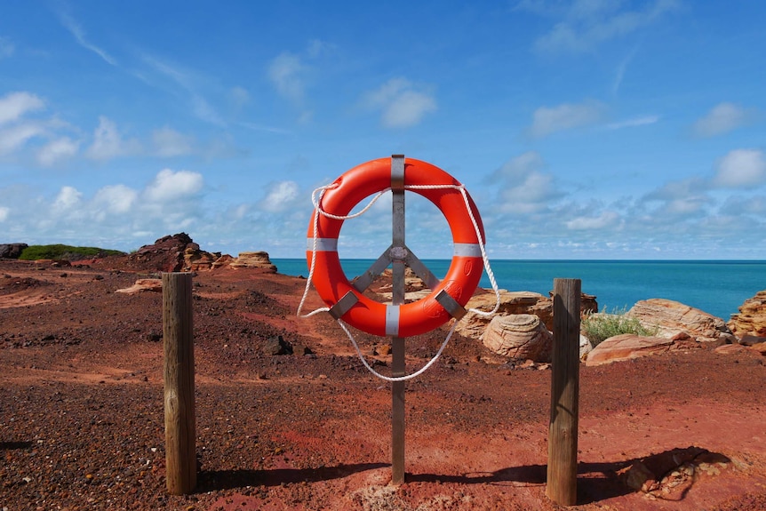 A life ring mounted on a steel pole in red dirt atop a cliff with the blue ocean behind it and clouds in the sky