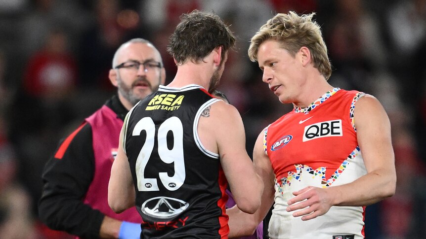 A Sydney Swans AFL player stands next to a St Kilda opponent who is being helped by a trainer, and speaks to the other player.