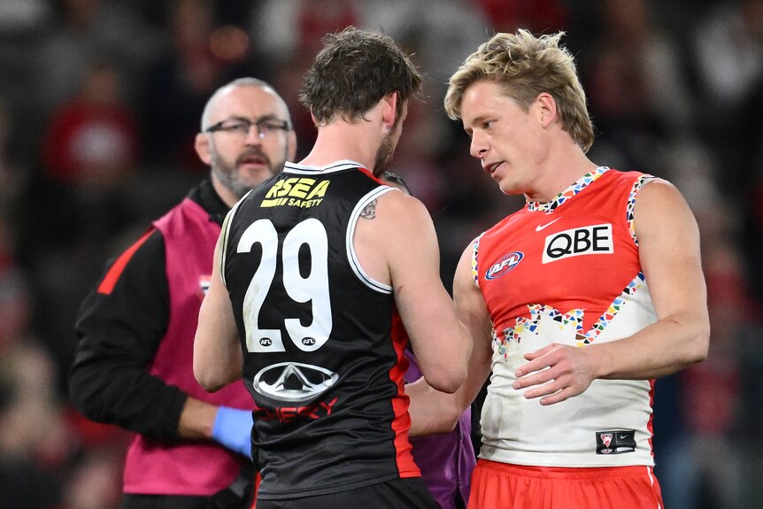 A Sydney Swans AFL player stands next to a St Kilda opponent who is being helped by a trainer, and speaks to the other player.