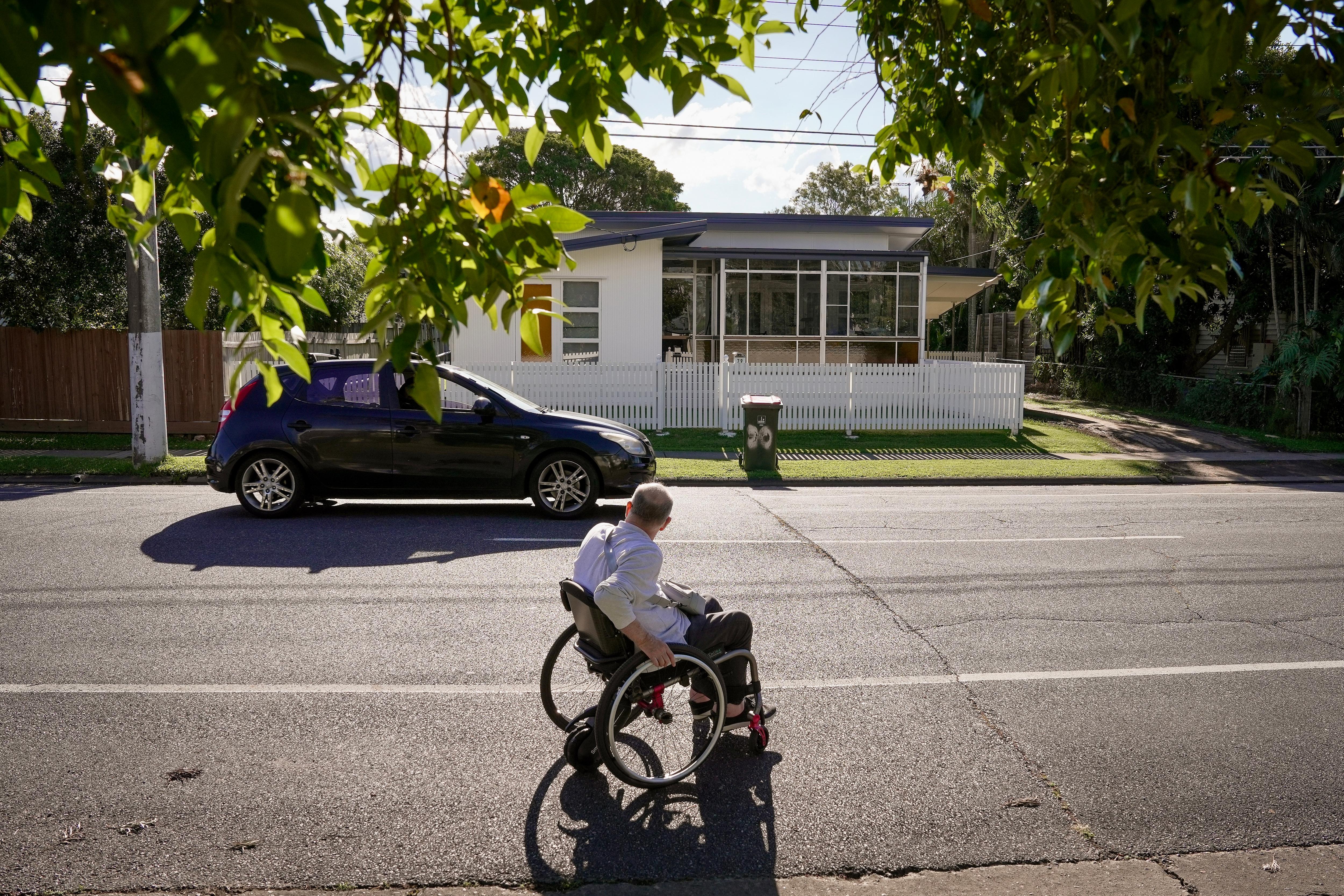 A middle aged white man with short grey hair. He's in a wheelchair, wheeling down a surbuban road