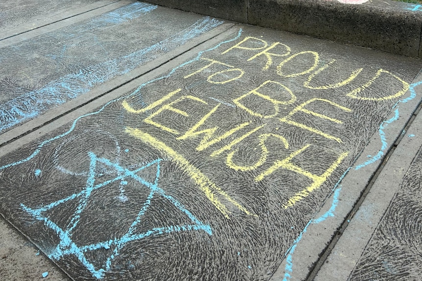 Yellow chalk on pavement saying 'Proud to be Jewish' with a Star of David drawn in blue chalk underneath