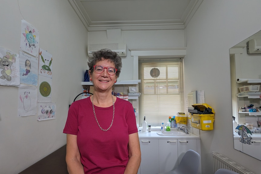 An older woman wearing a red top and glasses is pictured  in her GP consulting room. She is smiling.