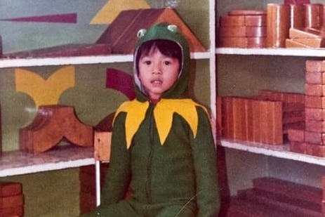 A young boy in a frog costume sitting on a stool in a classroom.