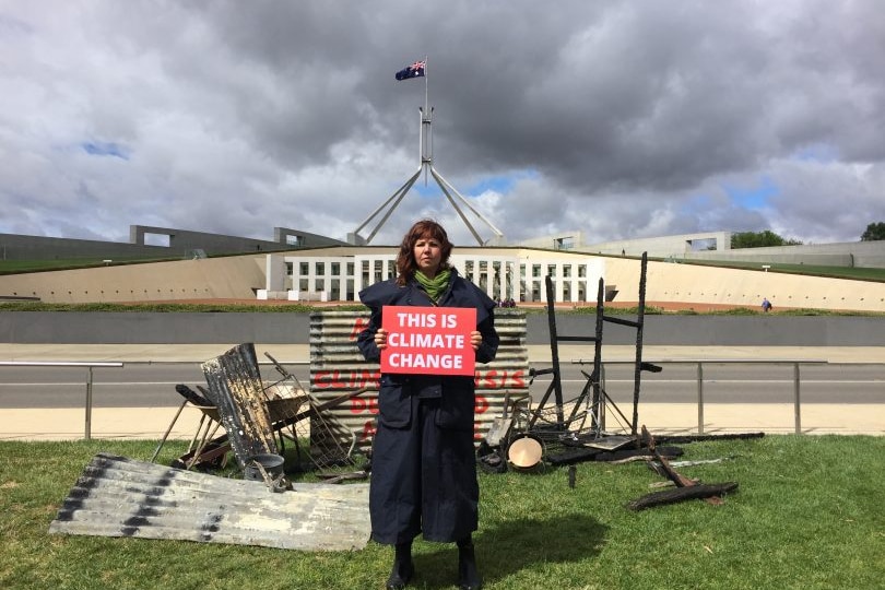 A woman stands in front of parts of burnt house on the lawn ofparliament house holding a sign that says this is climate change