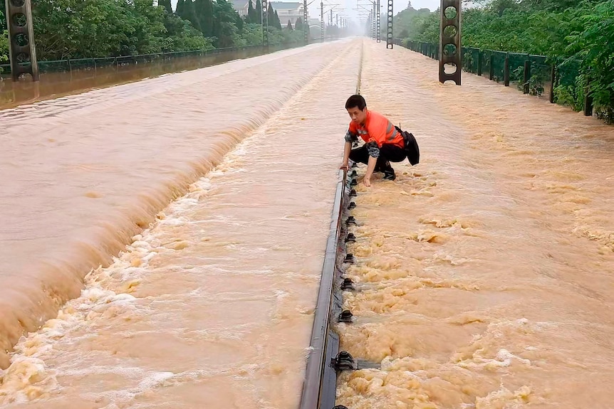 A man in hi-vis orange squats down at railway tracks that are almost covered in brown floodwater