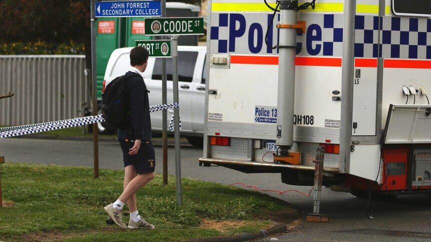 A boy in school uniform walks past a police car and a length of crime scene tape.
