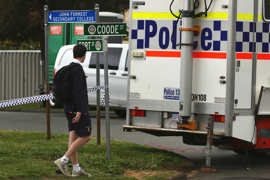 A boy in school uniform walks past a police car and a length of crime scene tape.