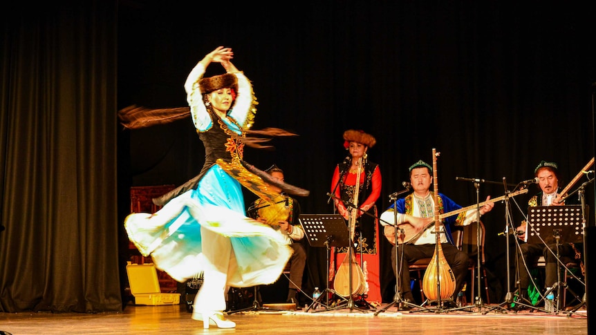 A Uyghur woman spins as she dances at a musical performance in Melbourne. Two men are playing the lute behind her on stage.